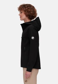 Zwarte softshell jas met een capuchon, voorzien van een ritsvak op de borst, zijzakken en een wit logo op de linker mouw.