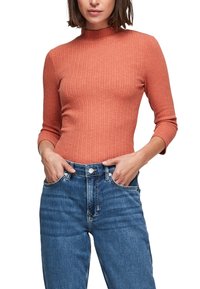 Gerippter, hochgeschlossenes Oberteil aus rostorangefarbener Stoff, mit langen Ärmeln. Kombiniert mit blauen, hoch geschnittenen Jeans aus Denim mit Vordertaschen.