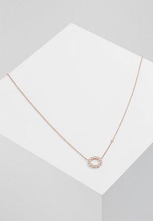 Collier en or rose avec une chaîne délicate et un pendentif circulaire orné de petites pierres claires, posé sur un présentoir géométrique blanc.
