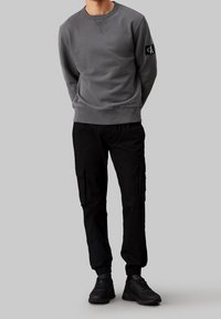 Grå sweatshirt med rund halsringning och ribbade muddar, tillsammans med svarta cargo-byxor med sidofickor och smalnande anklar. Svarta sneakers.