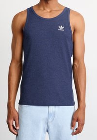 Camiseta sin mangas de rayas en color navy, hecha de algodón, con un cuello redondo y un pequeño logo blanco de Adidas en el lado izquierdo del pecho. Combinada con jeans de color azul claro.