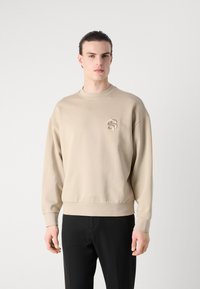 Beige színű pulóver, bordázott kerek nyakkal, ejtett vállakkal és egy kis hímzett logóval a mellkason. Sima anyag, laza fazon.