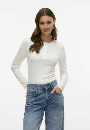 Haut blanc à manches longues avec une texture côtelée et un col rond, présentant une patte de boutonnage. Associé à un jean en denim bleu à coupe ample.