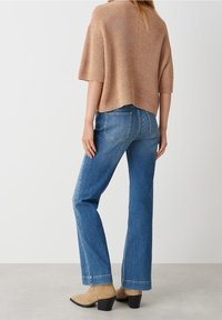 Brązowy dzianinowy oversize sweter z krótkim rękawem, jasnoniebieskie proste spodnie dżinsowe, beżowe botki za kostkę z grubym obcasem.