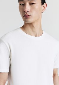 Giovane uomo con capelli neri corti che indossa una maglietta bianca a girocollo semplice, che guarda leggermente di lato su uno sfondo neutro.