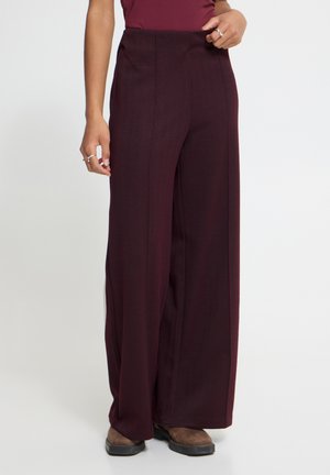 Personne portant un pantalon large taille haute violet foncé avec des coutures latérales, des chaussures marron et un haut foncé assorti sur un fond uni.