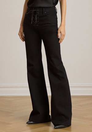 Donna che indossa pantaloni neri a vita alta e gamba larga con dettaglio anteriore stringato, abbinati a scarpe nere con punta, in piedi su pavimento di legno.