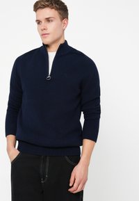 Pullover in blu navy lavorato a maglia con colletto alto e zip a quarto. Le maniche e l'orlo a costine aggiungono texture e struttura.