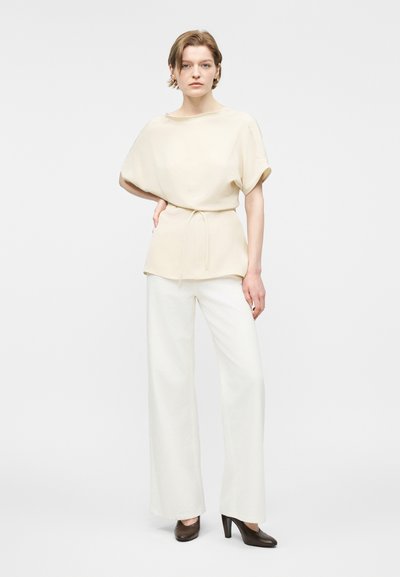 Femme debout, main sur la hanche, portant un haut crème à manches courtes avec ceinture à la taille, pantalon blanc à jambes larges, et chaussures à talons hauts marron.