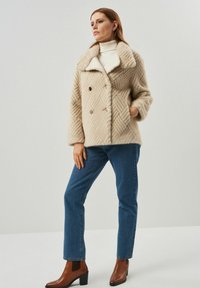 Beżowy dwurzędowy płaszcz z futra syntetycznego w diamentowy wzór, z szerokim kołnierzem i złotymi guzikami, w połączeniu z niebieskimi dżinsami i brązowymi botkami za kostkę.