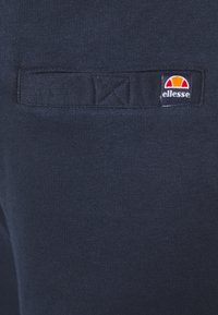 Navyblaue Sweatpants mit glatter Textur, die auf dem Bereich der hinteren Tasche ein kleines Logo-Patch in Rot, Gelb und Weiß aufweisen.
