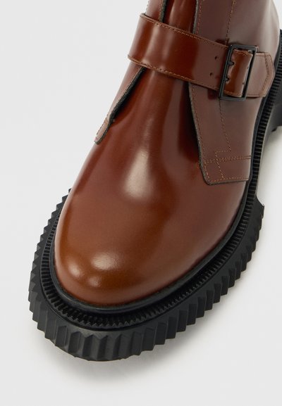 Καφέ δερμάτινο παπούτσι με μαύρη ραβδωτή ελαστική σόλα και λουρί με αγκράφα στο πλάι στο πάνω μέρος.