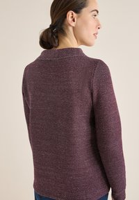 Kobieta z brązowymi włosami związanymi w kucyk, ubrana w teksturowany, długi rękaw maroon sweter, widoczna z tyłu na jednolitym beżowym tle.