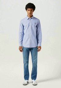 Camisa azul de botões com um pequeno logótipo bordado, combinada com jeans azul claro e ténis brancos com pormenores cinzentos.