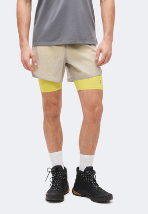 Muž nosi vrstvené béžové šortky cez žlté kompresné šortky, biele ponožky a čierne turistické topánky, stojí na svetlom pozadí.