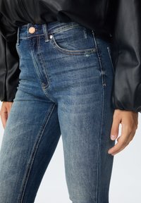 Mršavi traperice srednje plave boje s ispranim područjima i suptilnim oštećenjima. Imaju bakreni dugme i kontrastne šavove.