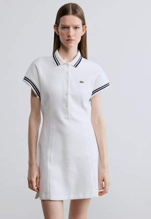 Witte polojurk met korte mouwen, voorzien van een marineblauw gestreepte kraag en een klein groen logo op de borst. Gladde textuur en getailleerde pasvorm.
