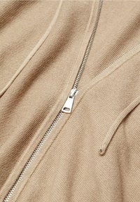 Gros plan sur un tissu beige en tricot avec une fermeture éclair en métal argenté et un cordon de serrage, montrant les détails de la texture et des coutures.