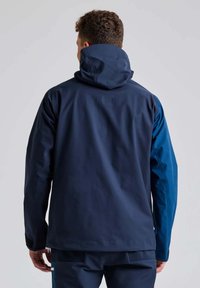 Giacca impermeabile blu navy con cappuccio regolabile, caratterizzata da un accento blu sulla manica sinistra e una texture liscia. Polsini regolabili.