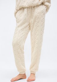Απαλά παντελόνια lounge από βελούδινο φλις σε κρεμ απόχρωση με υφή πλεκτού σχεδίου καλωδίου, ελαστικές μανσέτες και εμπρόσθιες τσέπες, φορεμένα ξυπόλητα.