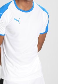 Man som bär en vit sporttröja med blå axlar och kanter, med en blå Puma-logotyp på bröstet.