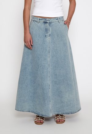 Persona che indossa una gonna lunga di jeans azzurri chiari, una maglietta bianca e sandali beige con decorazioni di perle, in piedi davanti a uno sfondo semplice.