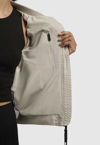 Jachetă bej ușoară cu manșete striate, interior texturat, buzunar cu fermoar și cordon de ajustare la margine pentru un fit personalizabil.