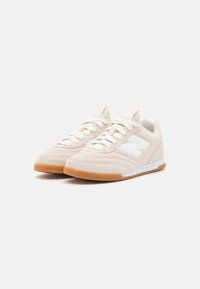 Un par de zapatillas de ante beige con cordones blancos, logo blanco en el lateral y suelas de goma color caramelo sobre un fondo blanco.