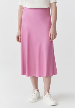 Vrouw met een midi-lange roze rok, witte top en witte sneakers, staand tegen een effen lichte achtergrond.