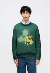 Zöld kötött pulóver nagy sárga Mickey egér grafikával az elején és fekete felirattal a bal ujjon. Bordázott nyak- és mandzsettarész.