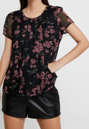 Femme portant un chemisier noir transparent à imprimé floral rose et un short en cuir noir, la main posée sur la hanche, sur un fond uni.