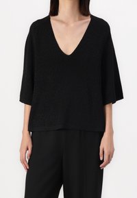 Femme portant un haut noir en maille à col en V ample avec des manches jusqu'au coude, assorti à un pantalon noir, sur un fond blanc uni.