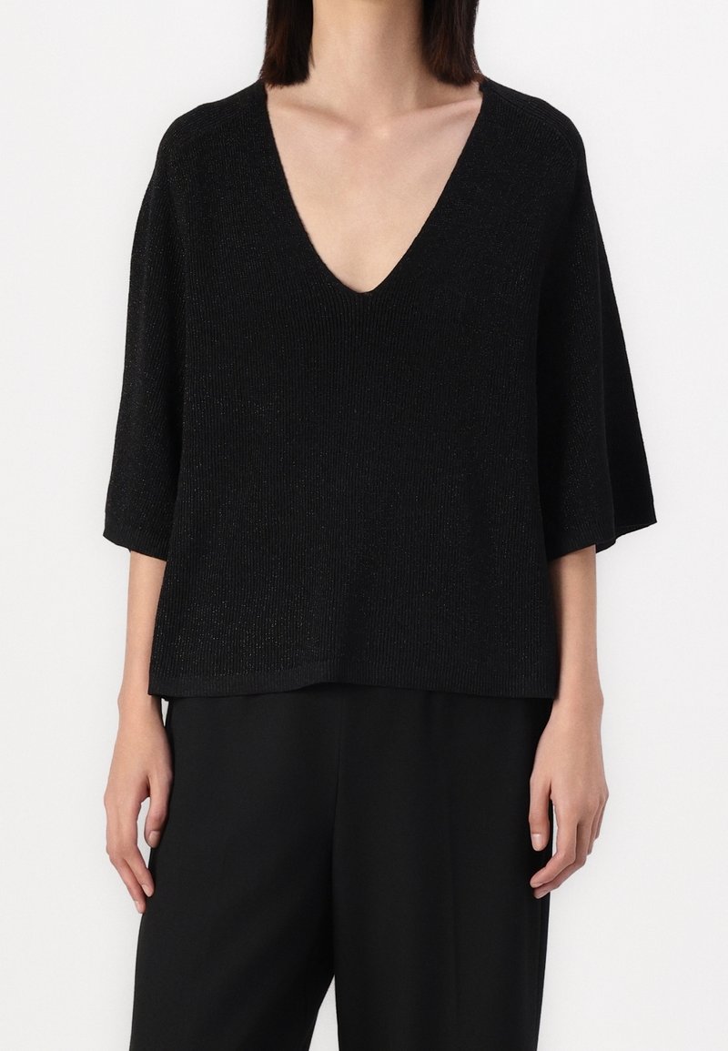 Femme portant un haut noir en maille à col en V ample avec des manches jusqu'au coude, assorti à un pantalon noir, sur un fond blanc uni.