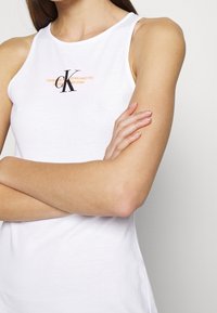 Bílý bezrukávový top vyrobený z měkké bavlny. Obsahuje malé černé logo CK s oranžovým textem. Jednoduchý, přiléhavý design.