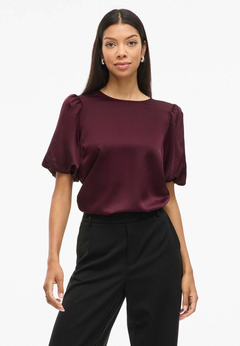 Blusa di satin bordeaux con maniche corte a sbuffo, scollatura rotonda e una texture liscia, abbinata a pantaloni neri su misura.