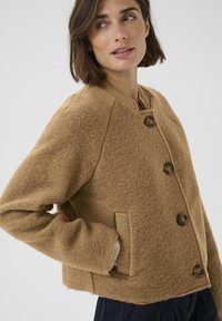 Giacca marrone in misto lana con collo tondo, quattro grandi bottoni e tasche laterali; presenta una forma squadrata e una texture morbida.