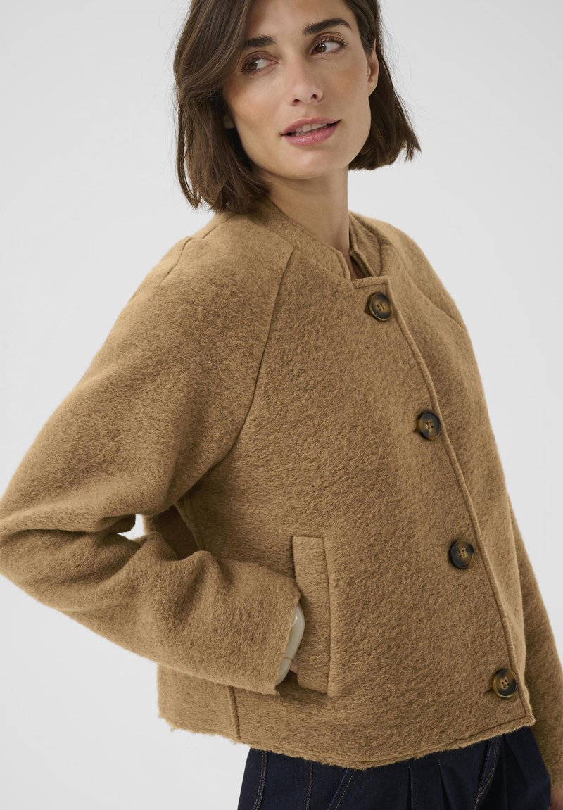 Giacca marrone in misto lana con collo tondo, quattro grandi bottoni e tasche laterali; presenta una forma squadrata e una texture morbida.