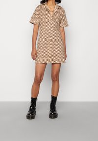 Robe à manches courtes en tissu beige avec un motif ondulé marron, boutonnée sur le devant avec un col, associée à des bottes de combat noires à lacets.