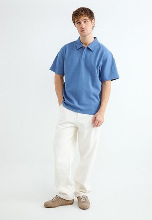 Jonge man staat met handen in de zakken, draagt een blauw poloshirt, witte broek en beige sneakers op een effen witte achtergrond.