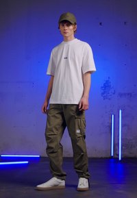 Weißes T-Shirt mit kurzen Ärmeln und kleinem blauen Logo, kombiniert mit olivfarbenen Cargo-Hosen mit Seitentaschen und weißen Turnschuhen.