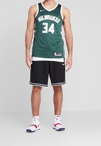 Canotta da basket verde con la scritta bianca "MILWAUKEE" e il numero "34", abbinata a pantaloni neri e sneakers bianche con dettagli rossi.