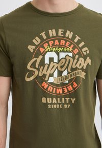 Maslinasto zelena pamučna majica s okruglim izrezom. Sadrži grafički dizajn s natpisima "Autentična Odjeća", "Superior" i "Kvaliteta od 87" u kontrastnim bojama.