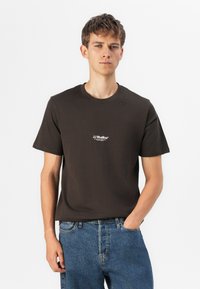 Camiseta de algodón marrón con cuello redondo y mangas cortas, con un pequeño logo blanco en el pecho. Combinada con pantalones vaqueros de mezclilla azul.