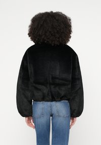 Άτομο με σγουρά μαλλιά, φορώντας μαύρο μαλακό μπουφάν και μπλε τζιν, με την πλάτη γυρισμένη προς ένα απλό λευκό φόντο.