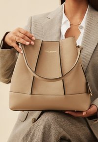 Sac à main en cuir beige au design structuré, avec une anse supérieure, un logo doré et des accents noirs contrastants. Texture lisse et intérieur spacieux.