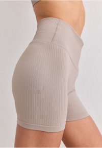 Gros plan d'une personne portant un short de cycliste beige côtelé taille haute, en position debout sur un fond clair uni.