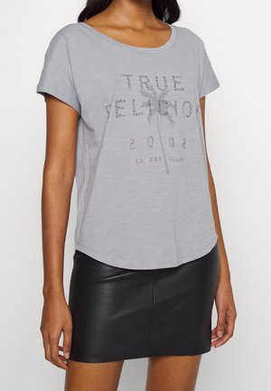 Frau trägt graues True Religion T-Shirt mit Palmenprint und schwarzen Leder-Minirock, steht vor weißem Hintergrund.