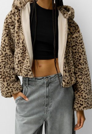 Persona che indossa una giacca corta in pelliccia sintetica con stampa leopardata sopra un top nero corto e jeans grigi larghi con una mano in tasca.
