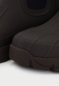 Acercamiento de botas de goma de textura marrón oscuro con suelas gruesas sobre una superficie blanca.