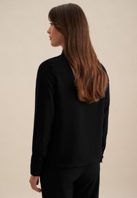 Zwarte blouse met lange mouwen, een gladde textuur, knoopmanchetten en een licht gebogen zoom, gezien vanaf de achterkant.